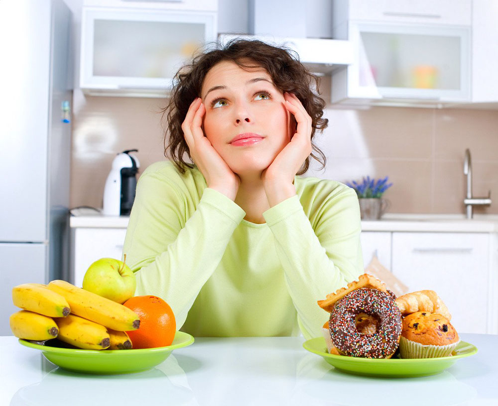 Woman in kitchen deciding between fruits and baked goods for breakfast