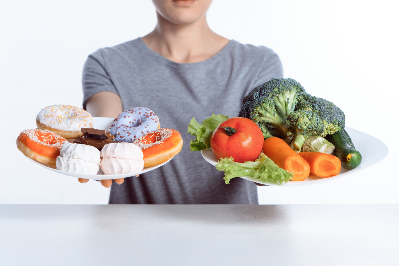 Woman holding a plate of donuts and high-carb foods in one hand and a plate of vegetables in the other hand.