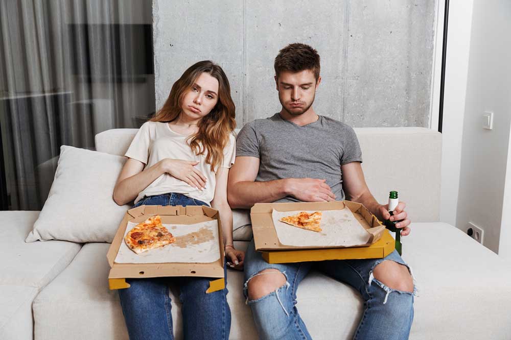 A couple sits on a couch feeling too full after overeating pizza.
