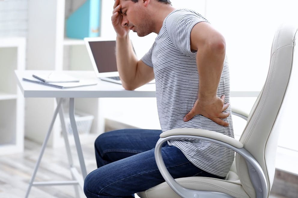 A man in a striped shirt at a desk holds hand to back and head, in pain, headache, back pain.
