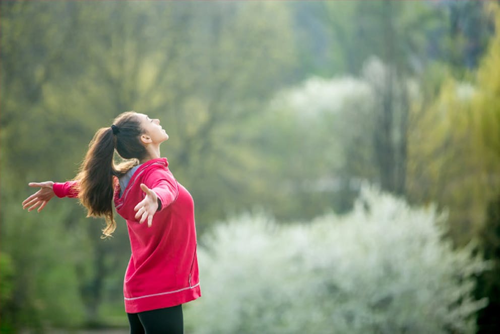 Young woman in red sweatshirt holds arms out to side, eyes closed, relaxing in nature outdoors.