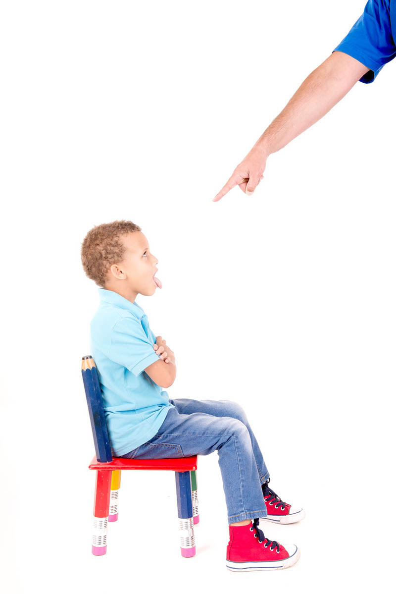 A frustrated young boy sits on a classroom chair, getting in trouble from adult pointing finger