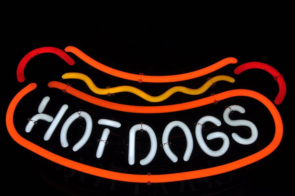 Neon sign with text and shape of hot dog, mustard, and bun – neon orange, yellow, and white.