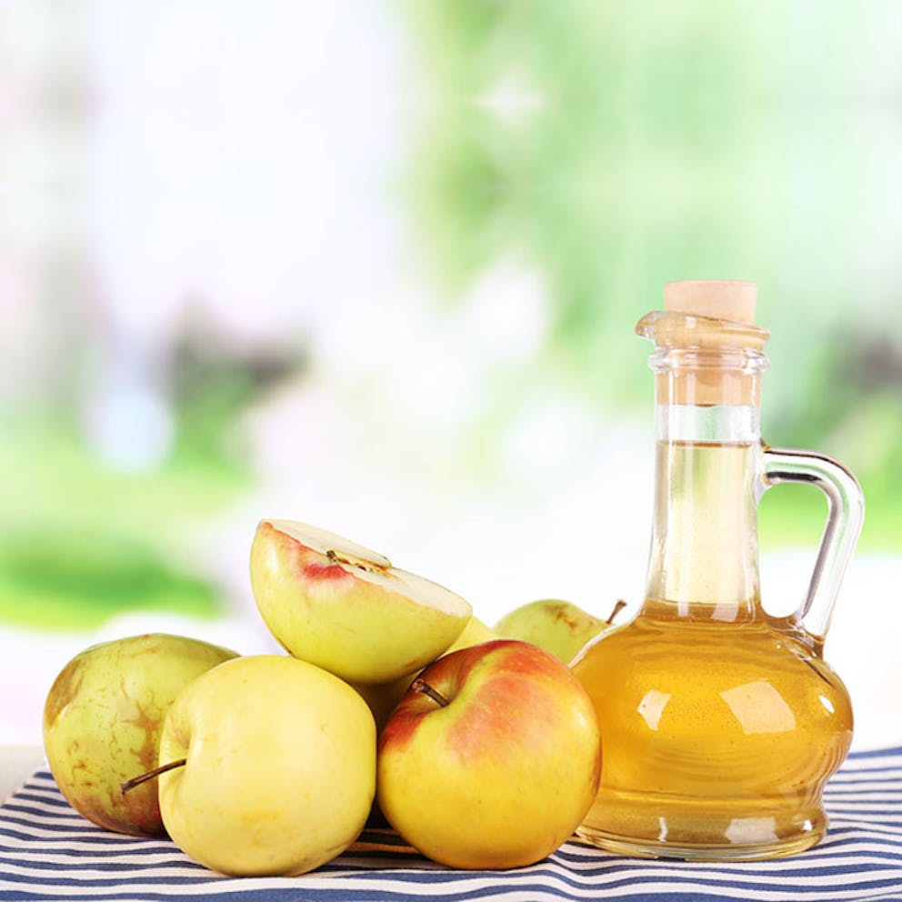 Pile of fresh apples next to glass bottle of apple cider vinegar on a striped cloth.