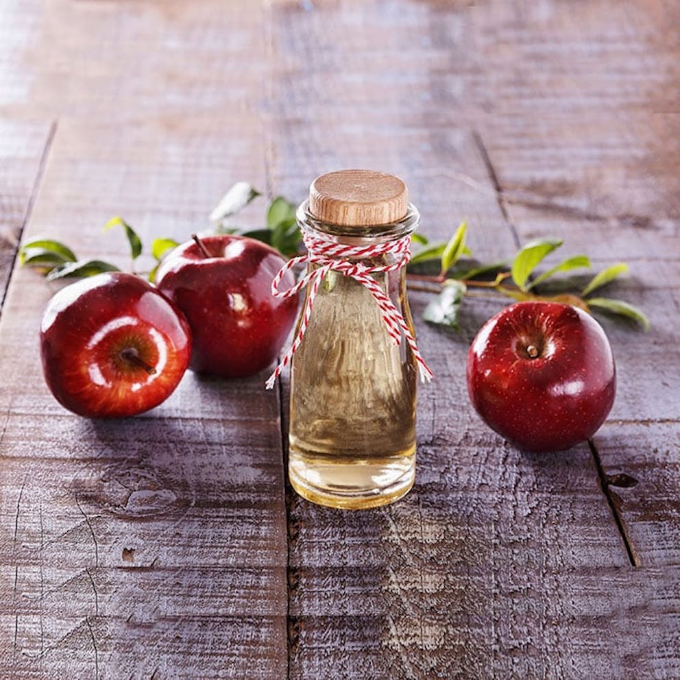Glass jar of apple cider vinegar with twine next to red apples and branches on rustic wooden surface.