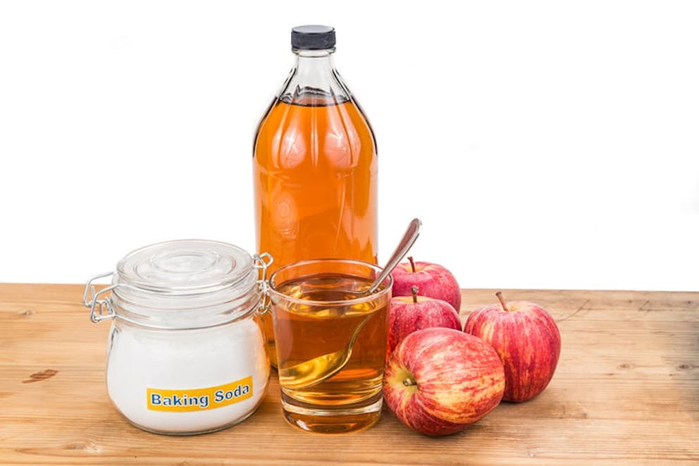 Top view of apples, apple cider vinegar bottle and glass, baking soda in jar, and spoon on table.