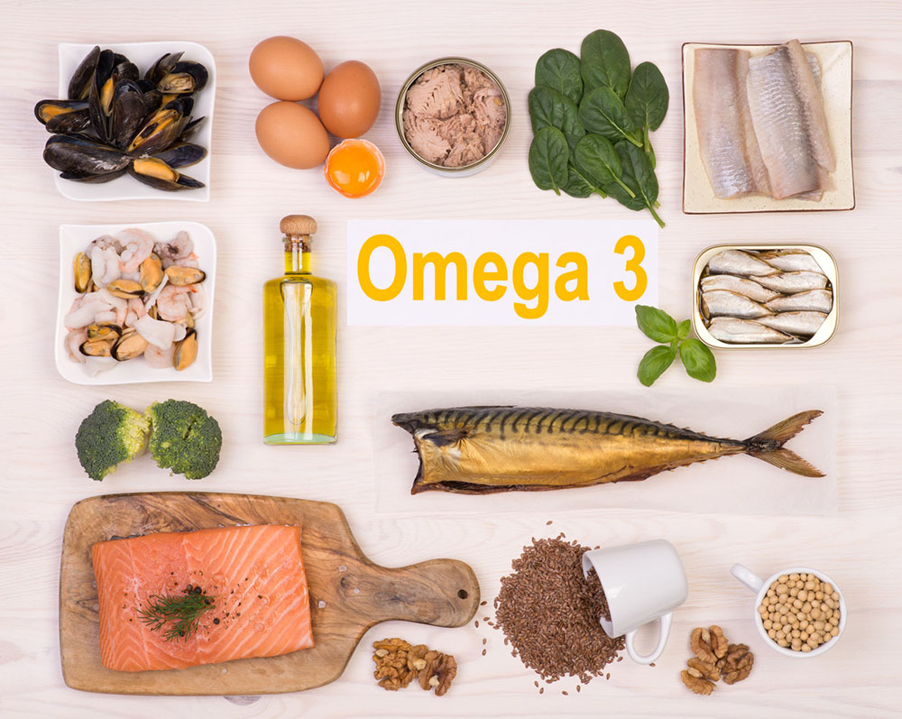 Foods high in omega 3s on a white table, including salmon, fish, flax, walnuts, eggs, and sardines.