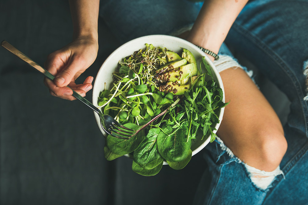 Girl in jeans holds fork and bowl of greens like spinach, arugula, sprouts, and avocado
