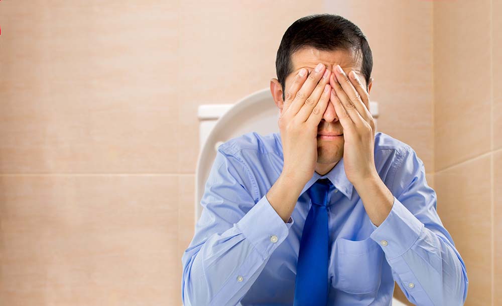 A businessman sits on the toilet with hands in his face, in pain from hemorrhoids.