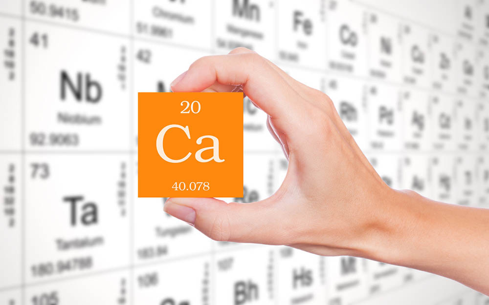 A hand holds the symbol for calcium on an orange square with the periodic table in the background.