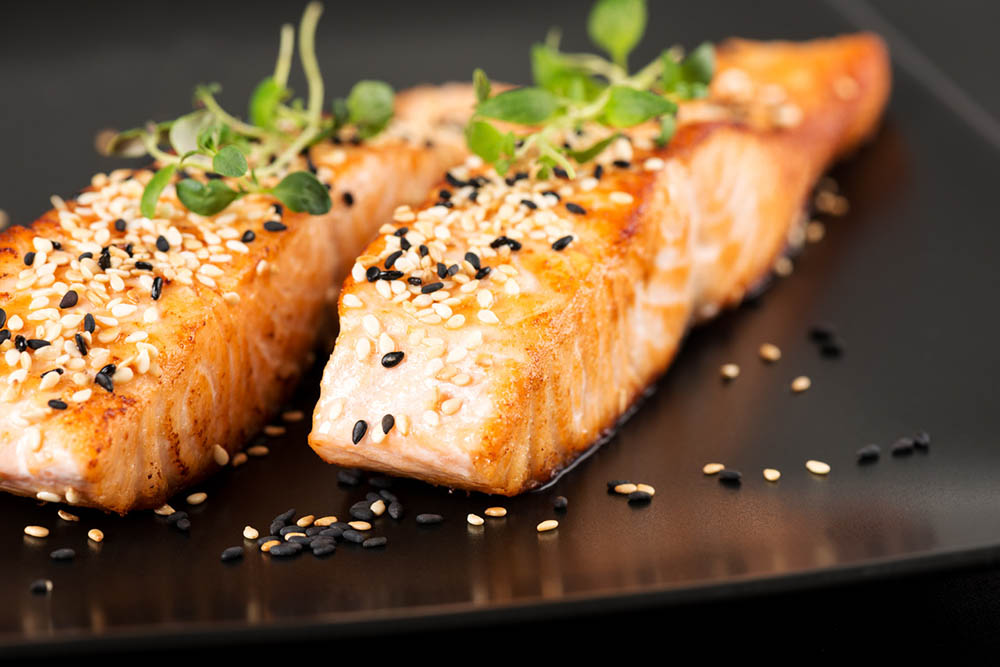 Grilled salmon topped with black and white sesame seeds on a black plate.