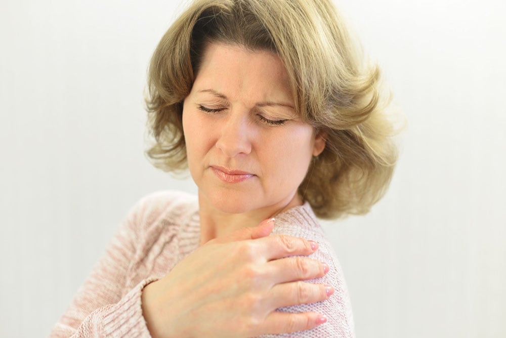 A woman with upper back and shoulder pain holds her hand to her shoulder and grimaces