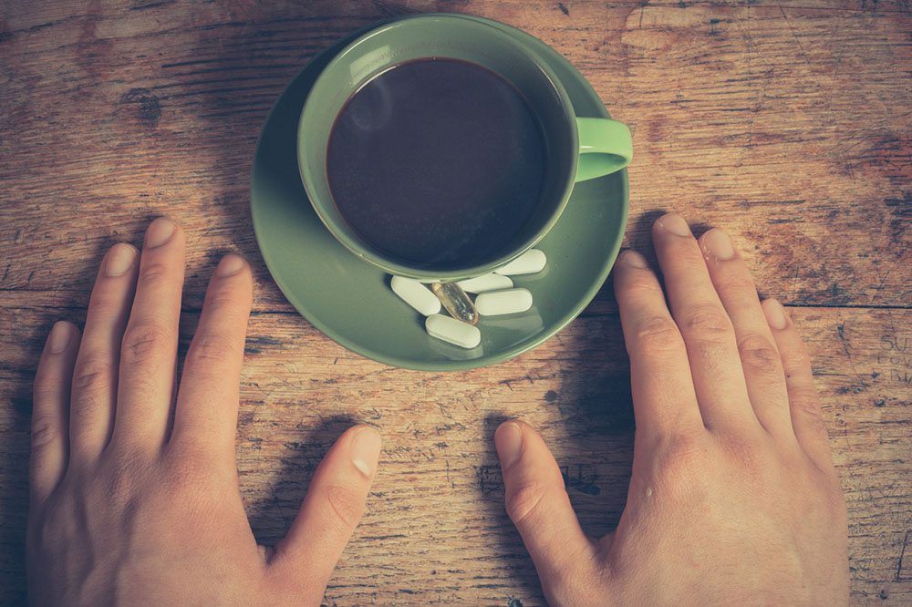 A person resting his hands next to a morning cup of coffee and vitamins.