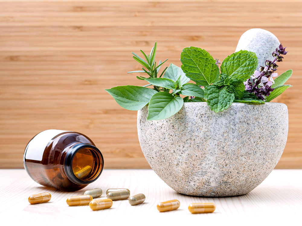 A bottle of herbal supplements spills out next to a mortar and pestle holding fresh herbs.