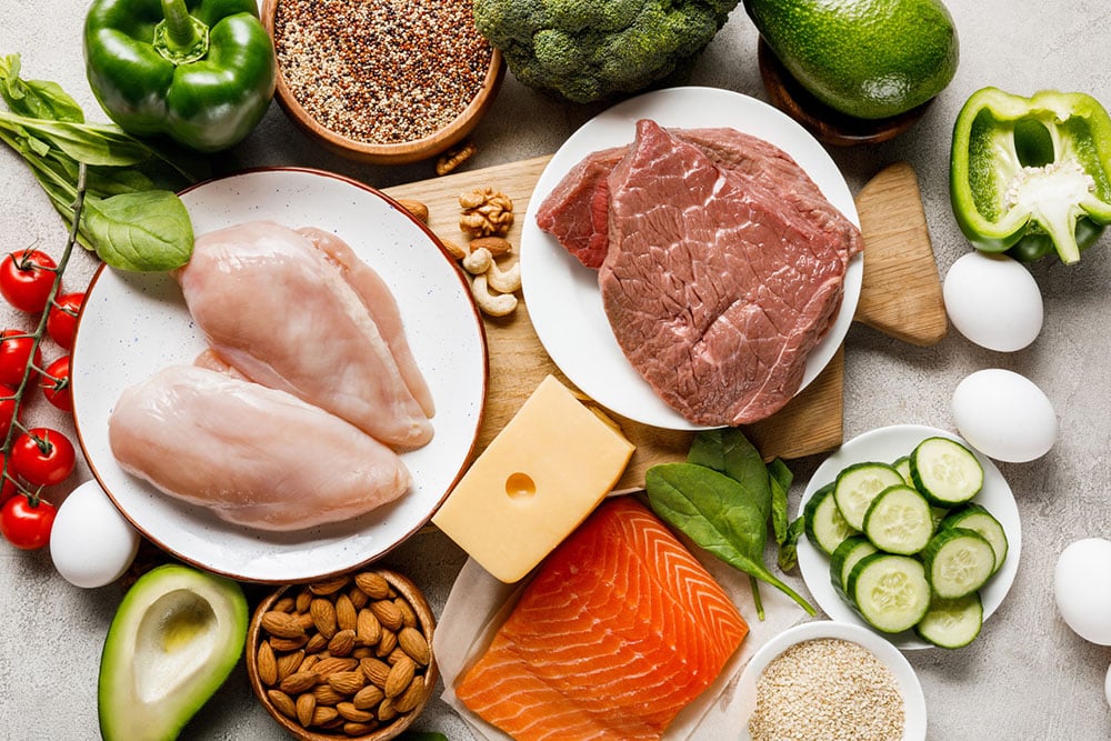 Ketogenic diet foods on a wood cutting board - salmon, chicken, avocados, eggs, and vegetables.