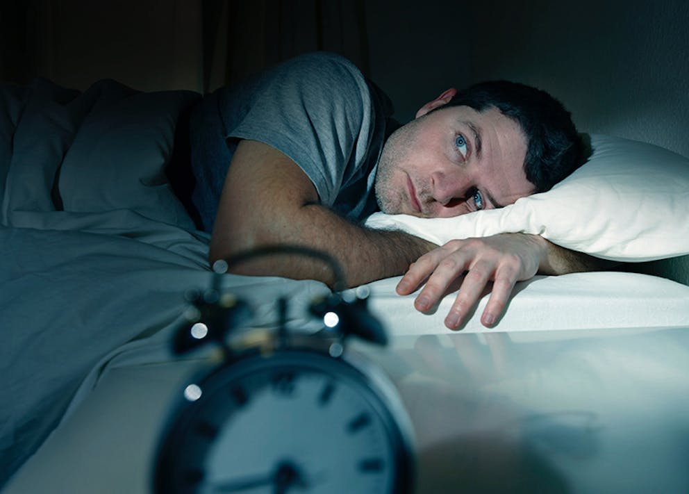 Man with insomnia lies in bed with eyes open next to alarm clock.