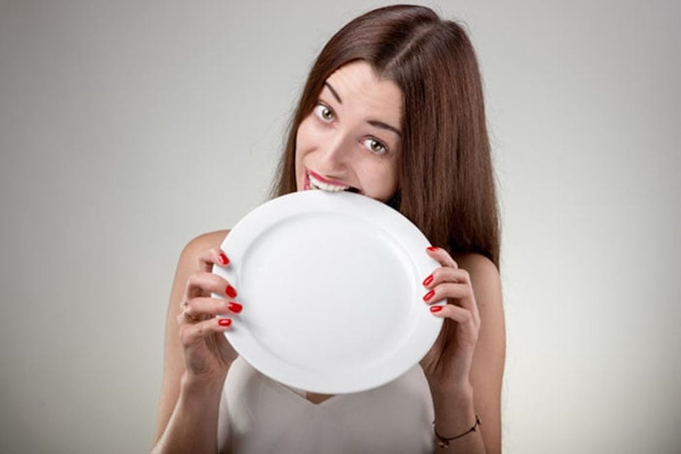 Hungry young woman holds white plate and bites on it, showing hunger and cravings for food.