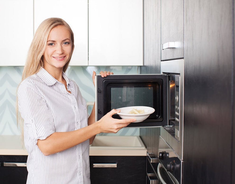 A young woman in white shirt puts white bowl of food into microwave oven.