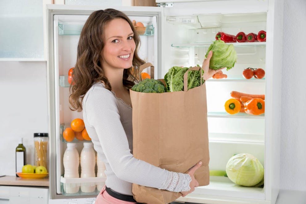 Happy Woman Holding Grocery Shopping Bag With Vegetables In Front Of Open Refrigerator | How To Stop Snacking Out Of Boredom | night time snacking