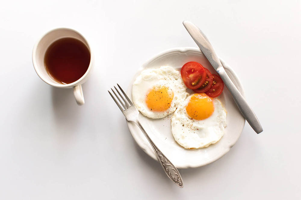 A breakfast meal of fried eggs on a plate with silverware next to a cup of coffee.