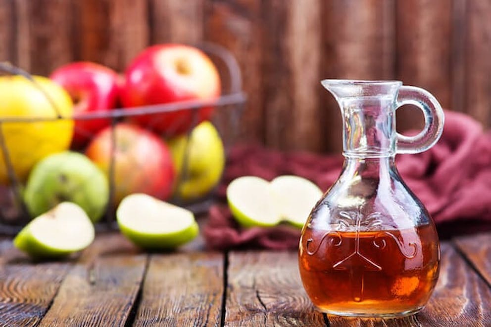 A pretty glass bottle of apple cider vinegar sitting on a wooden surface in front of a basket of apples | Gaining Weight During Lockdown? Do This!