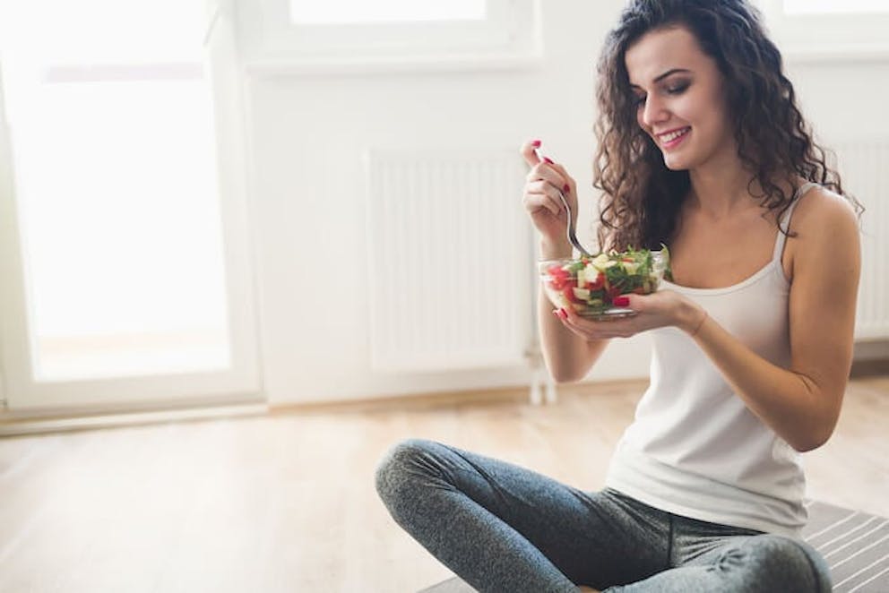 A healthy smiling woman sitting on the floor in exercise clothes eating a bowl of fresh salad | Gaining Weight During Lockdown? Do This!