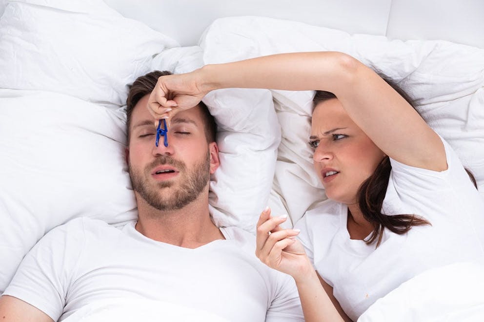 A frustrated woman in bed pinches husband’s nose with clip to make spouse stop snoring.