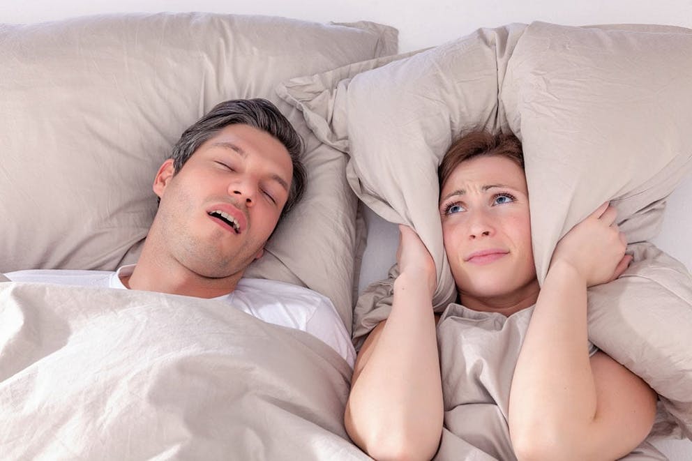 An annoyed woman holds her pillow over her ears next to sleeping snoring husband in bed.