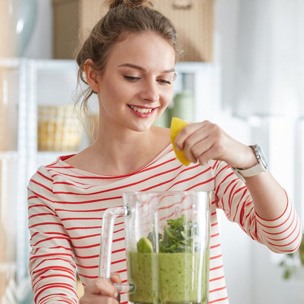 A smiling woman in a striped shirt squeezing lemon into a blender making a green kale shake.