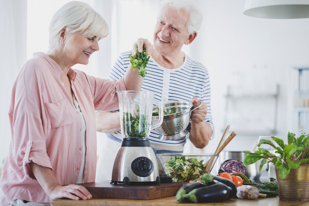 Elderly couple smiling while putting kale into a blender making a healthy green kale shake.