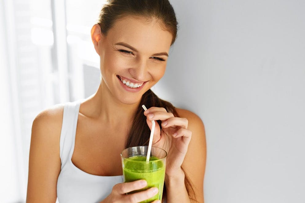 A smiling woman drinking a fresh green kale shake smoothie in a glass with a straw.