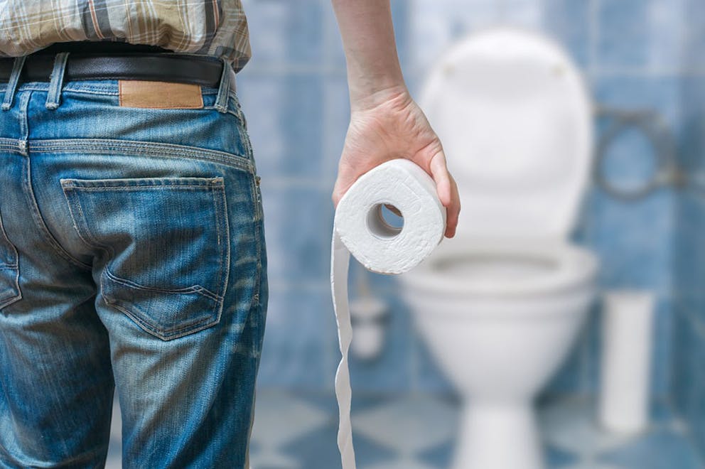 Man in jeans holds toilet paper in his hand while looking at a toilet, has constipation.