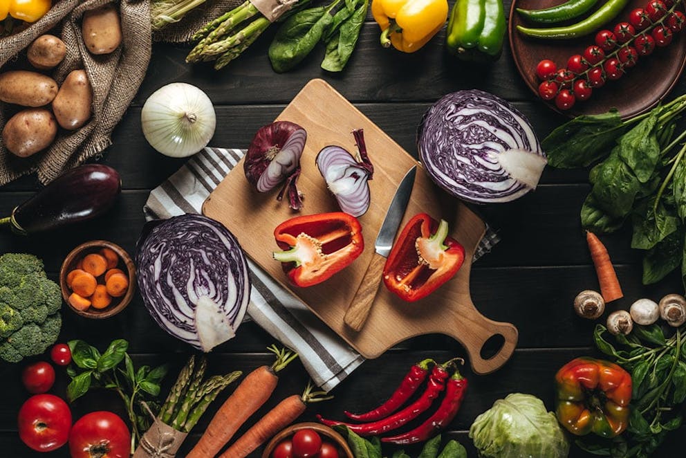 Assortment of fresh, healthy vegetables on and around wood cutting board with knife on dark table.
