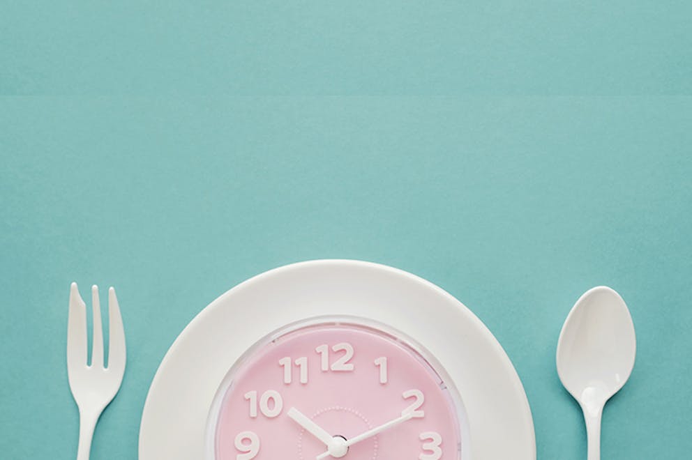 Intermittent fasting concept, white plate with pink clock on teal background, silverware.