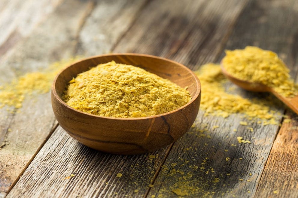 Wooden bowl full of yellow nutritional yeast, spoonful in background on wooden table.