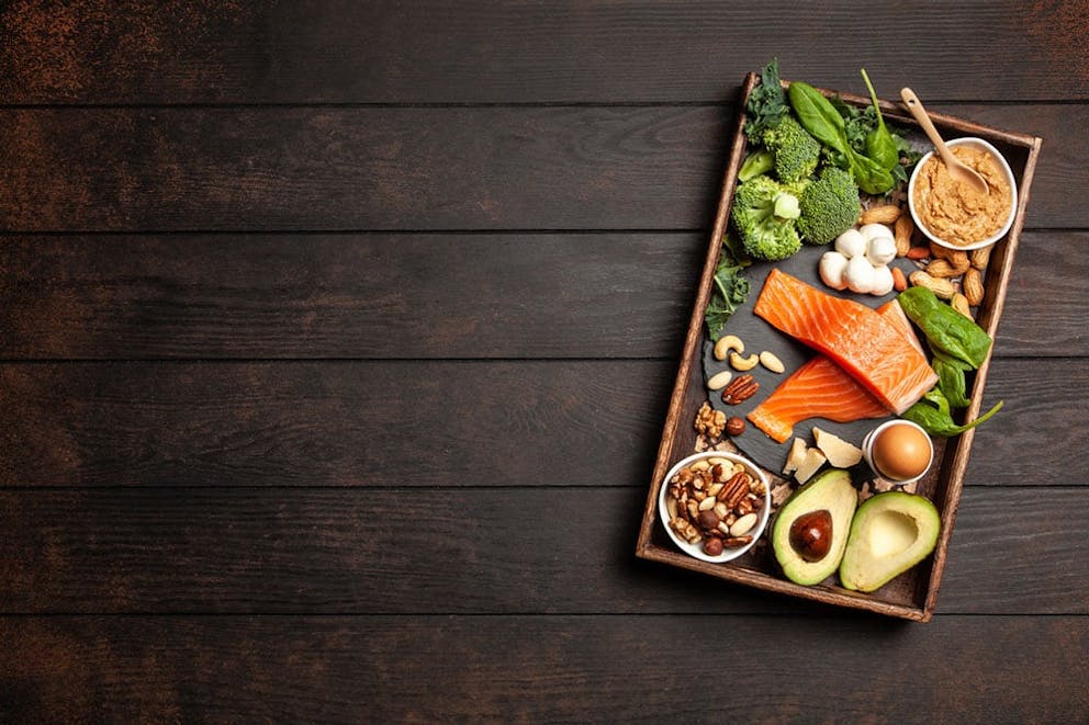 Healthy ketogenic diet foods on wooden tray on wood table. Fish, avocados, greens, nuts, egg.