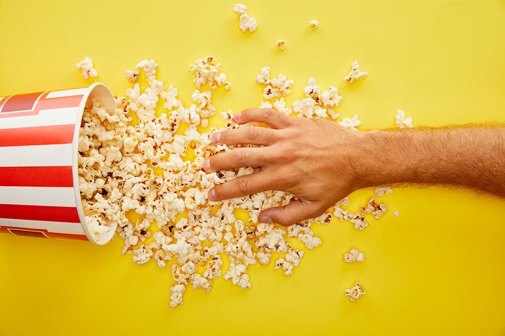 Hand grabbing popcorn spilling out of red and white movie popcorn bucket on yellow background.