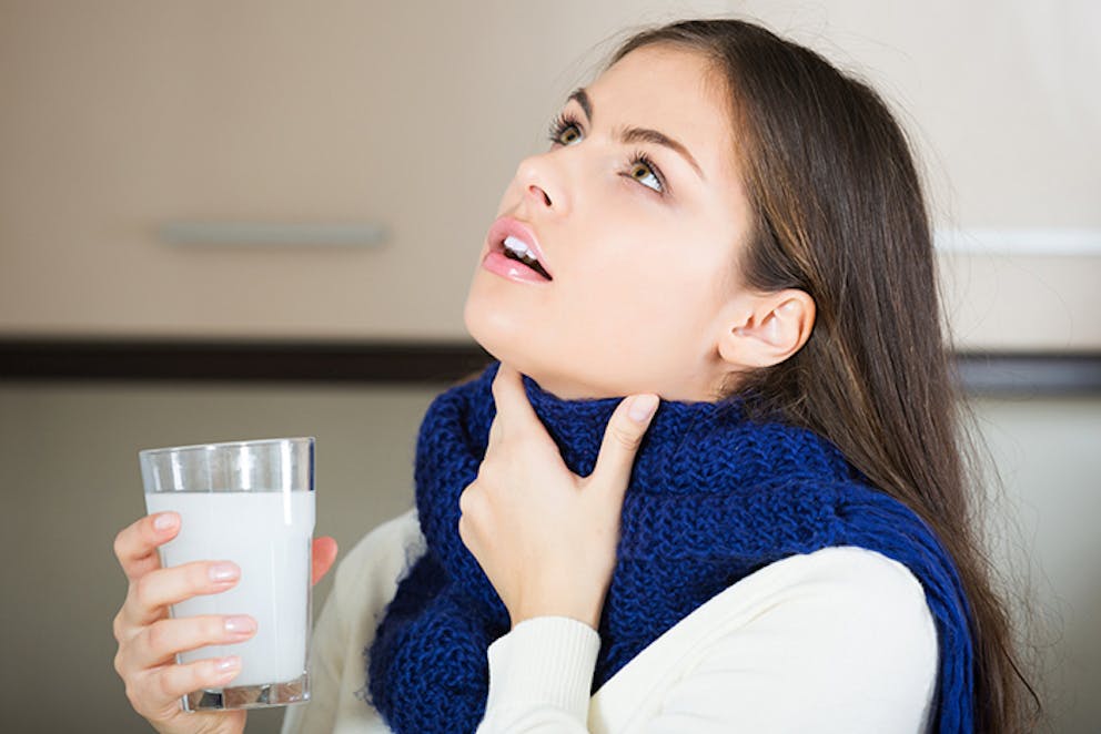 Young woman gargling wearing blue scarf holds hand to throat and glass cup, salt water gargle.