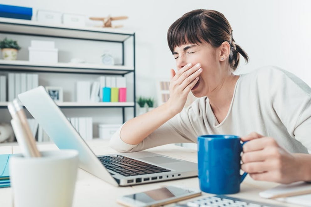 A tired woman yawns in her office in front of computer with coffee, sleep deprivation effects.