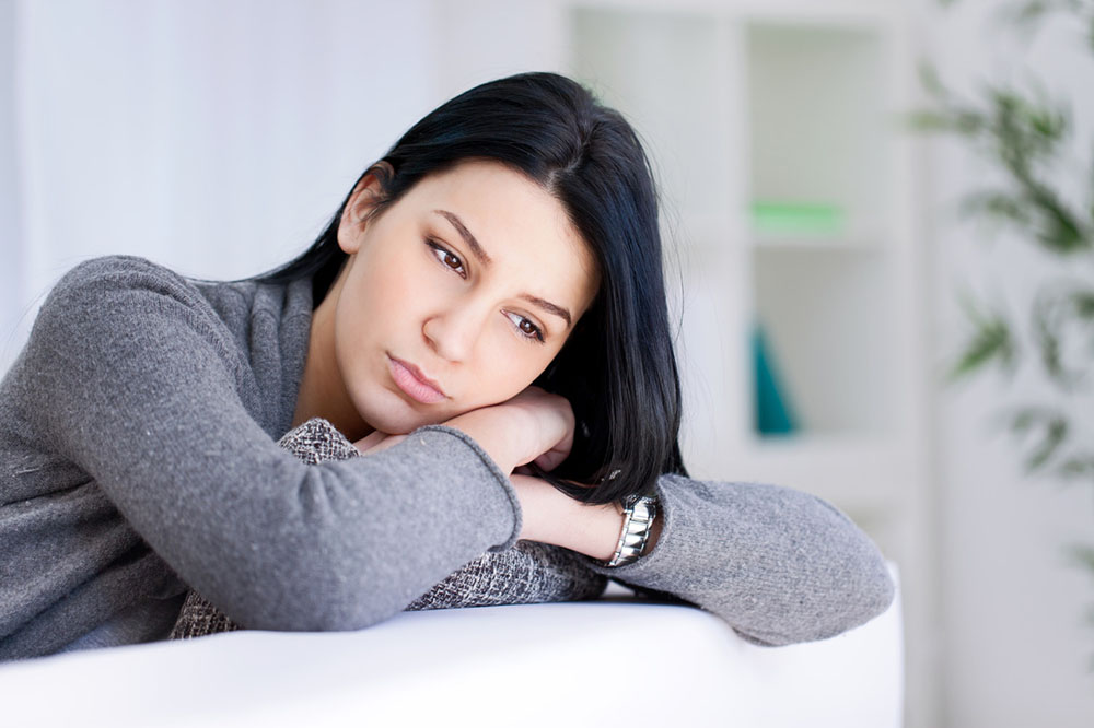 A depressed woman in a grey sweater leans on a white couch looking sad and feeling down.