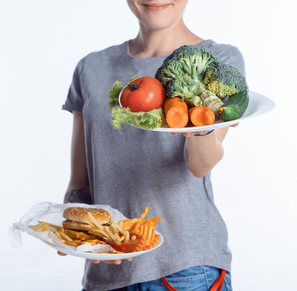 Woman holds a plate of healthy vegetables over an unhealthy plate of burger and fries.