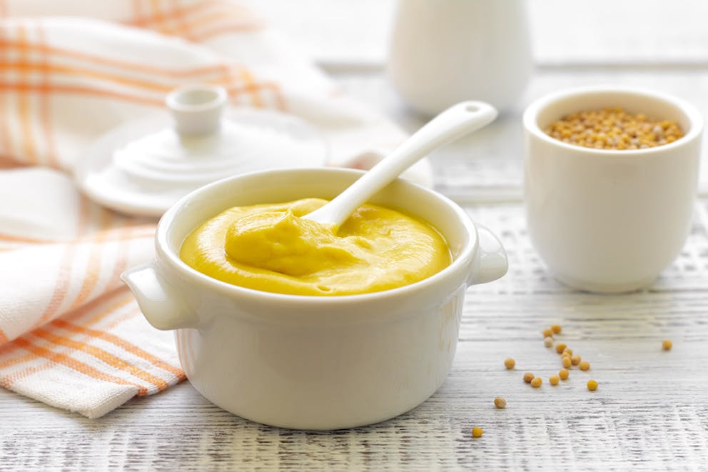 Mustard condiment in white dish with spoon, mustard seeds in background on wood table with cloth.