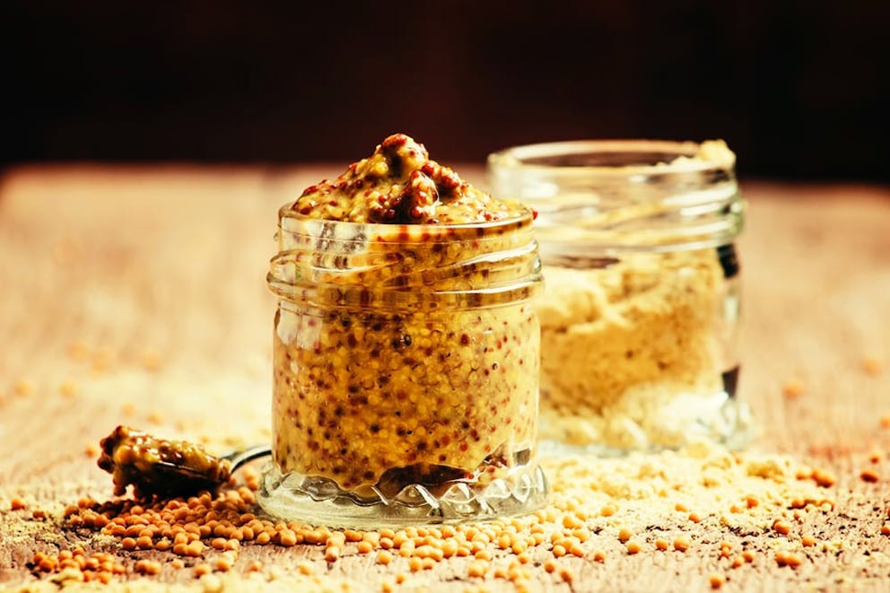 Whole grain mustard in small glass jars on table with mustard seeds and spices around it.