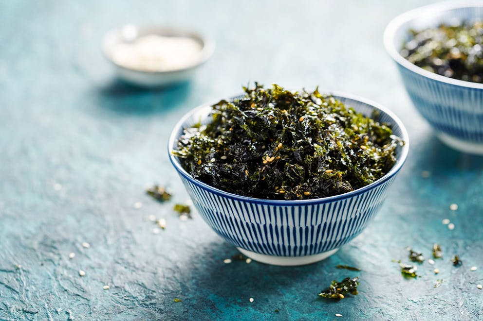 Crumbled salted roasted seaweed in blue and white bowl with sesame seeds and bowls in background.