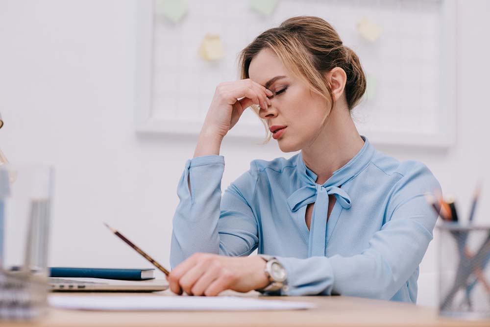 A stressed businesswoman sits at her office desk with a hand on her forehead.