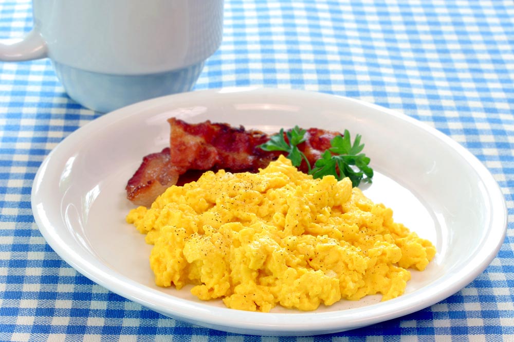 A plate of scrambled eggs and bacon on a blue checkered tablecloth