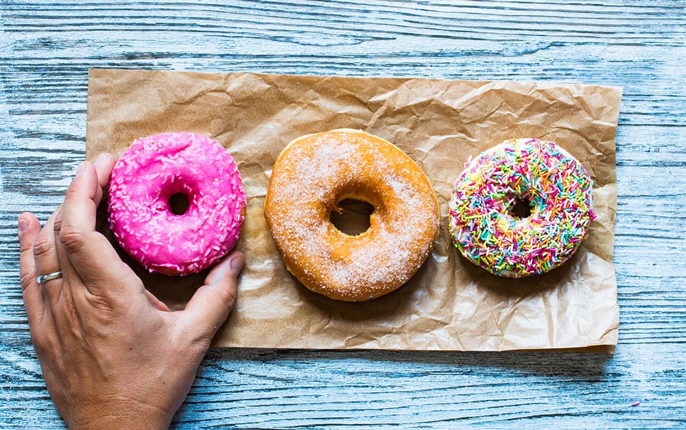 Brown paper on a blue wood table with three colorful donuts on top and a hand grabbing one donut.