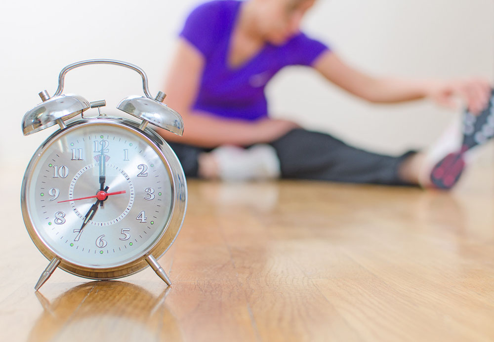 An old-fashioned alarm clock with a woman in workout clothes stretching in the background.