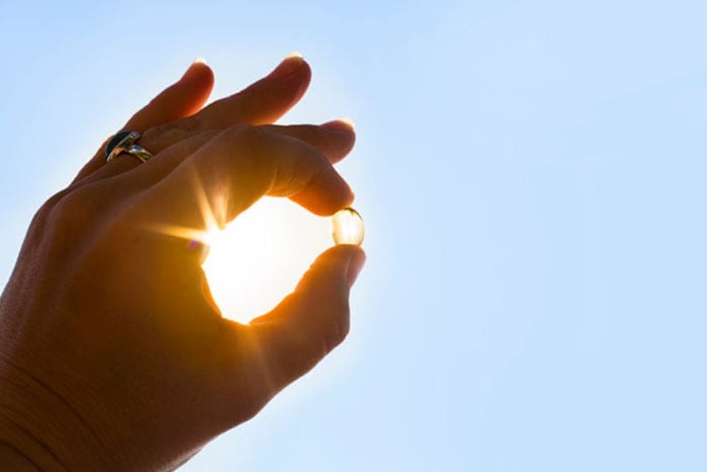 Hand holds vitamin D supplement pill with sunshine coming through, blue sky and sun exposure.