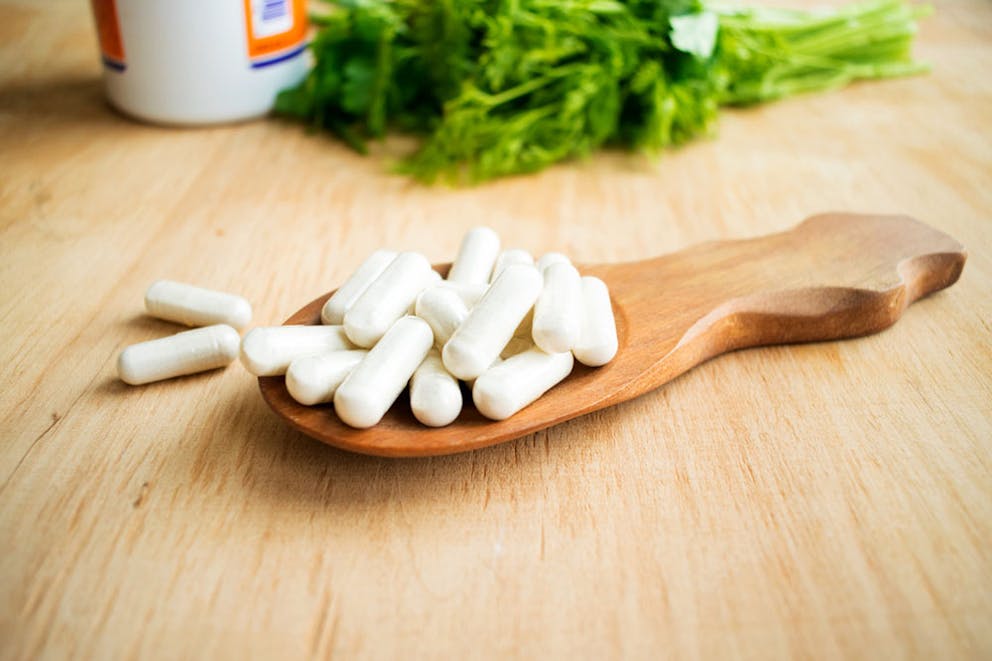 Capsules of vitamin K2 supplement on wooden spoon on a table with pill bottle and green herbs.